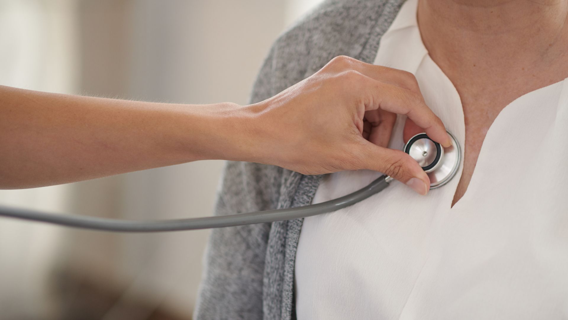 Plano aproximado da mão de uma pessoa a usar um estetoscópio para auscultar o peito de uma mulher.