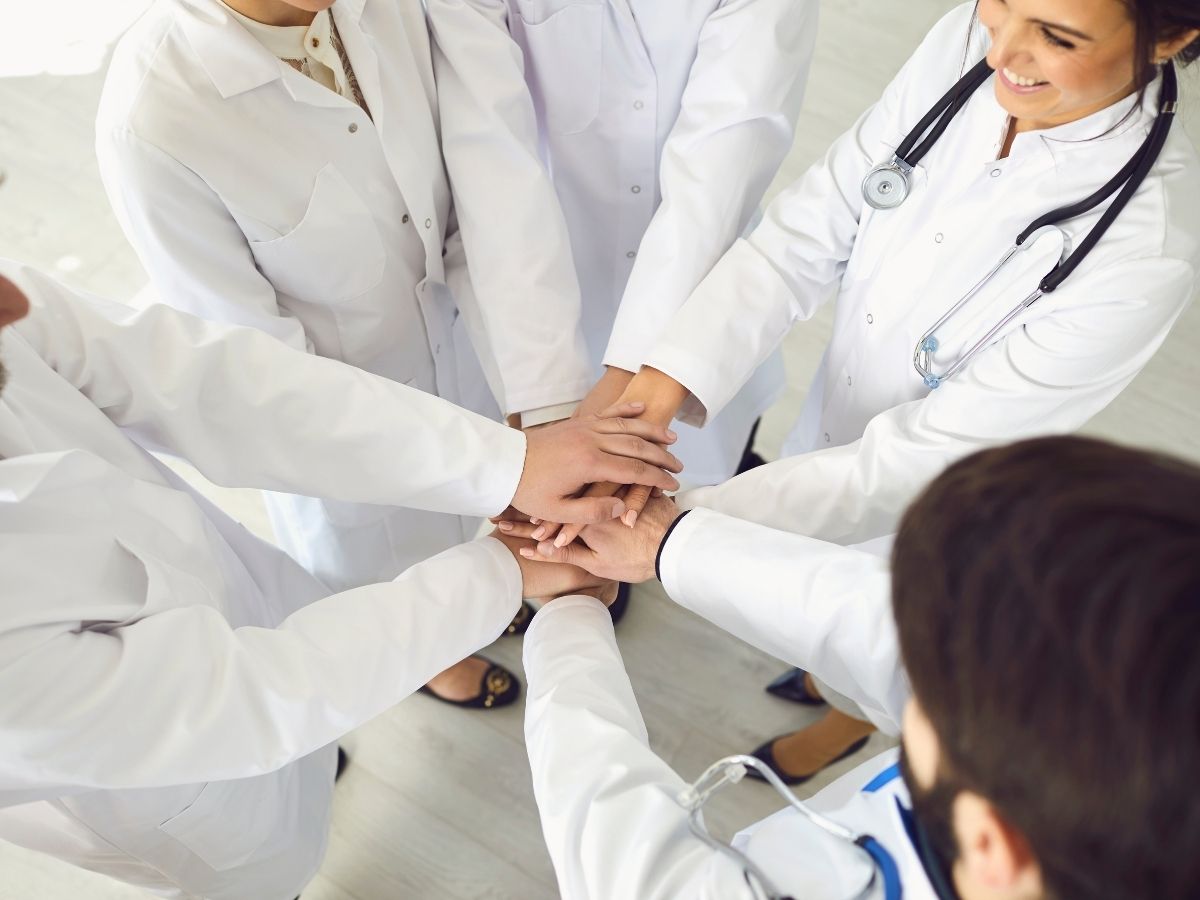 EVista de cima de um grupo de profissionais de saúde com batas brancas, a unir as mãos em conjunto num gesto de trabalho de equipa e união.