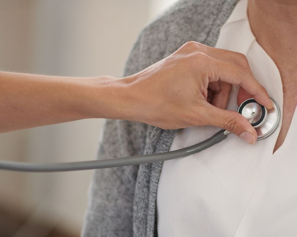 Plano aproximado da mão de uma pessoa a usar um estetoscópio para auscultar o peito de outra pessoa, que veste uma camisola branca e um casaco cinzento.