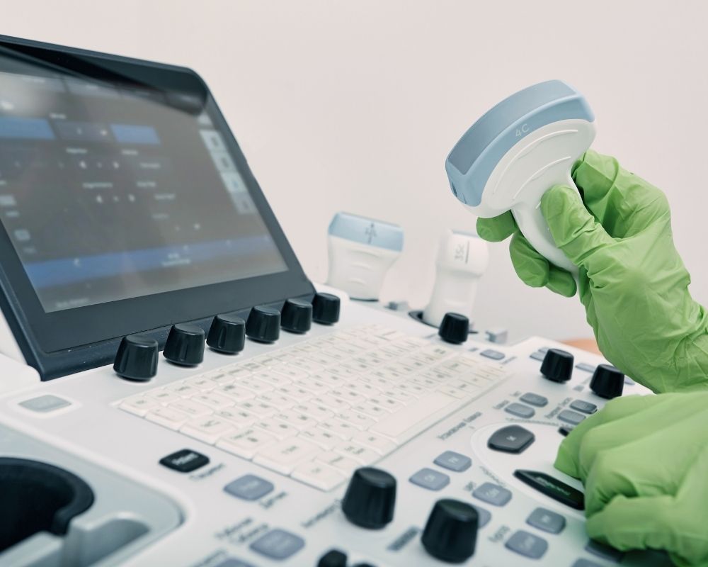 Plano aproximado de uma máquina de ecografia (ultrassom). Uma mão com luva verde manipula o transdutor do aparelho sobre o teclado e ecrã.