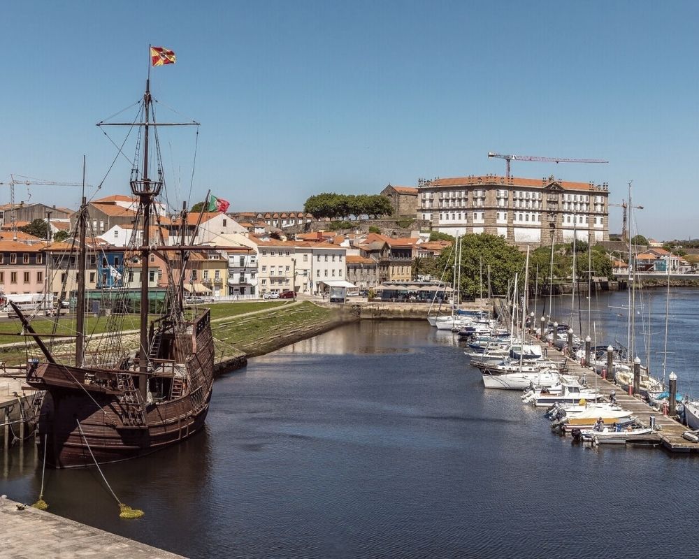 Vista de uma marina em Vila do Conde, com um navio de réplica de época atracado em primeiro plano, e o edifício do Convento de Santa Clara (ou semelhante) na margem.