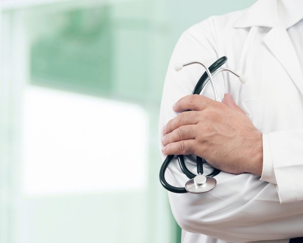 Plano aproximado de um médico com bata branca, de braços cruzados, a segurar um estetoscópio. O fundo é desfocado em tons de verde e branco.