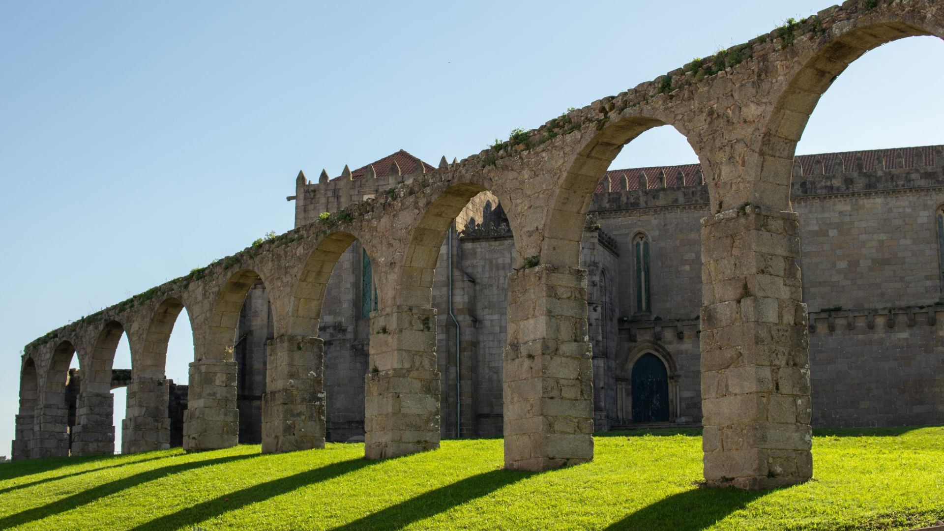 Arco de um antigo aqueduto (Arcos de São Francisco em Vila do Conde) em pedra, com uma igreja ou mosteiro antigo por trás, sob um céu azul, e relva verde em primeiro plano.